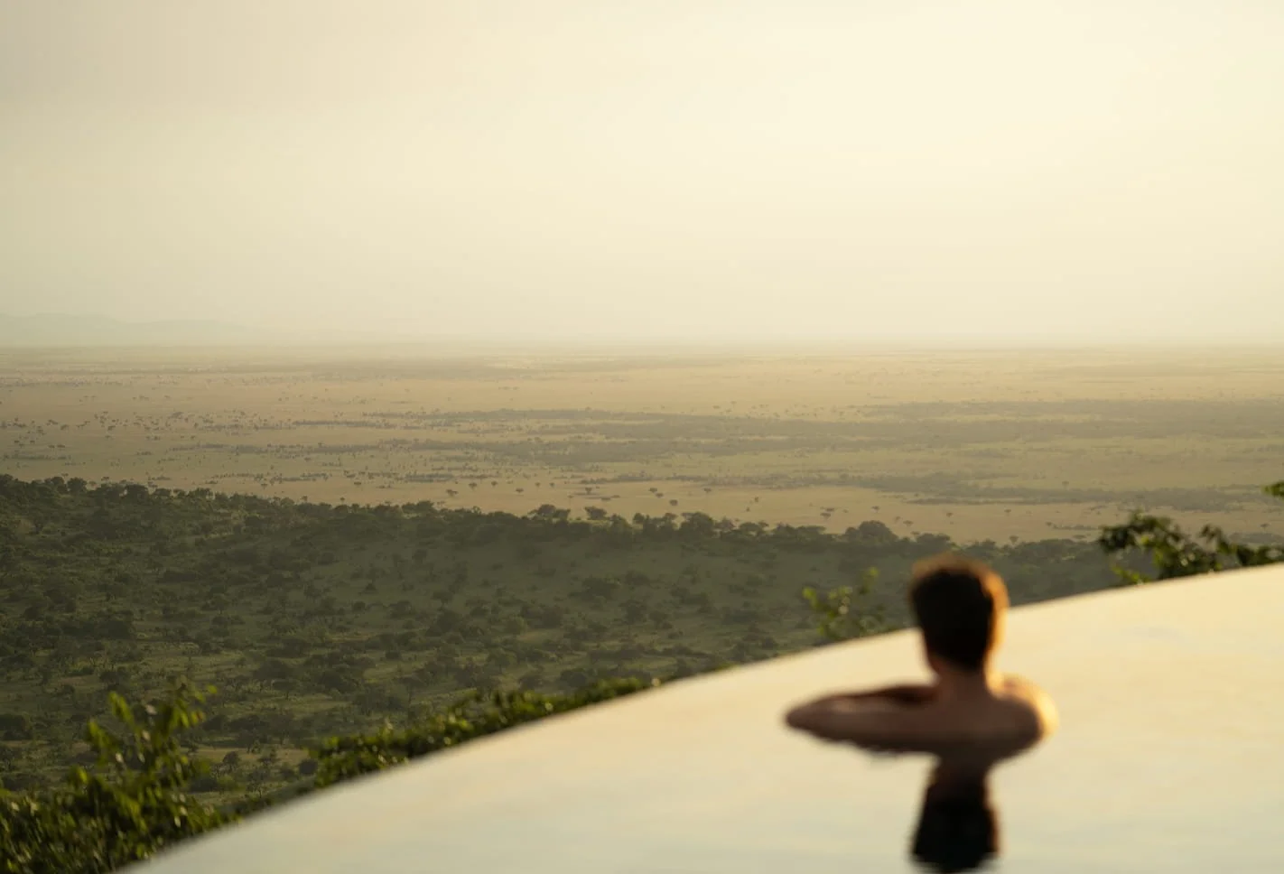 Person relaxing in an infinity pool overlooking a vast, flat landscape at sunset.