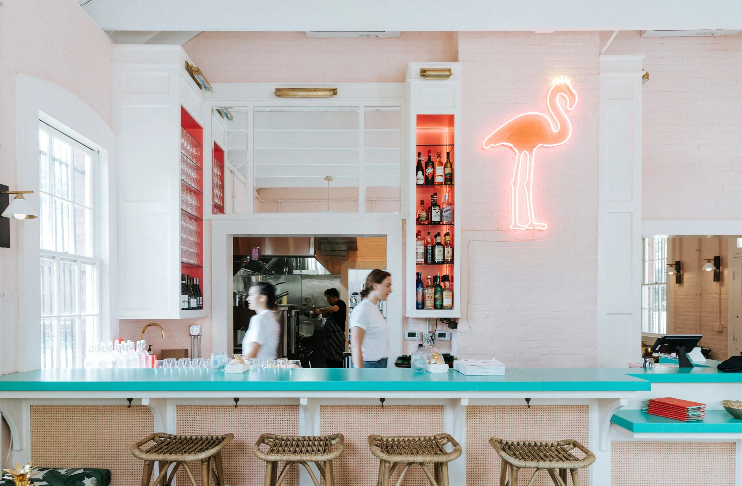 Inside a modern cafe with a pink and white color scheme. A neon flamingo sign on the wall, shelves with liquor bottles, kitchen area with staff, and a teal counter with glasses.