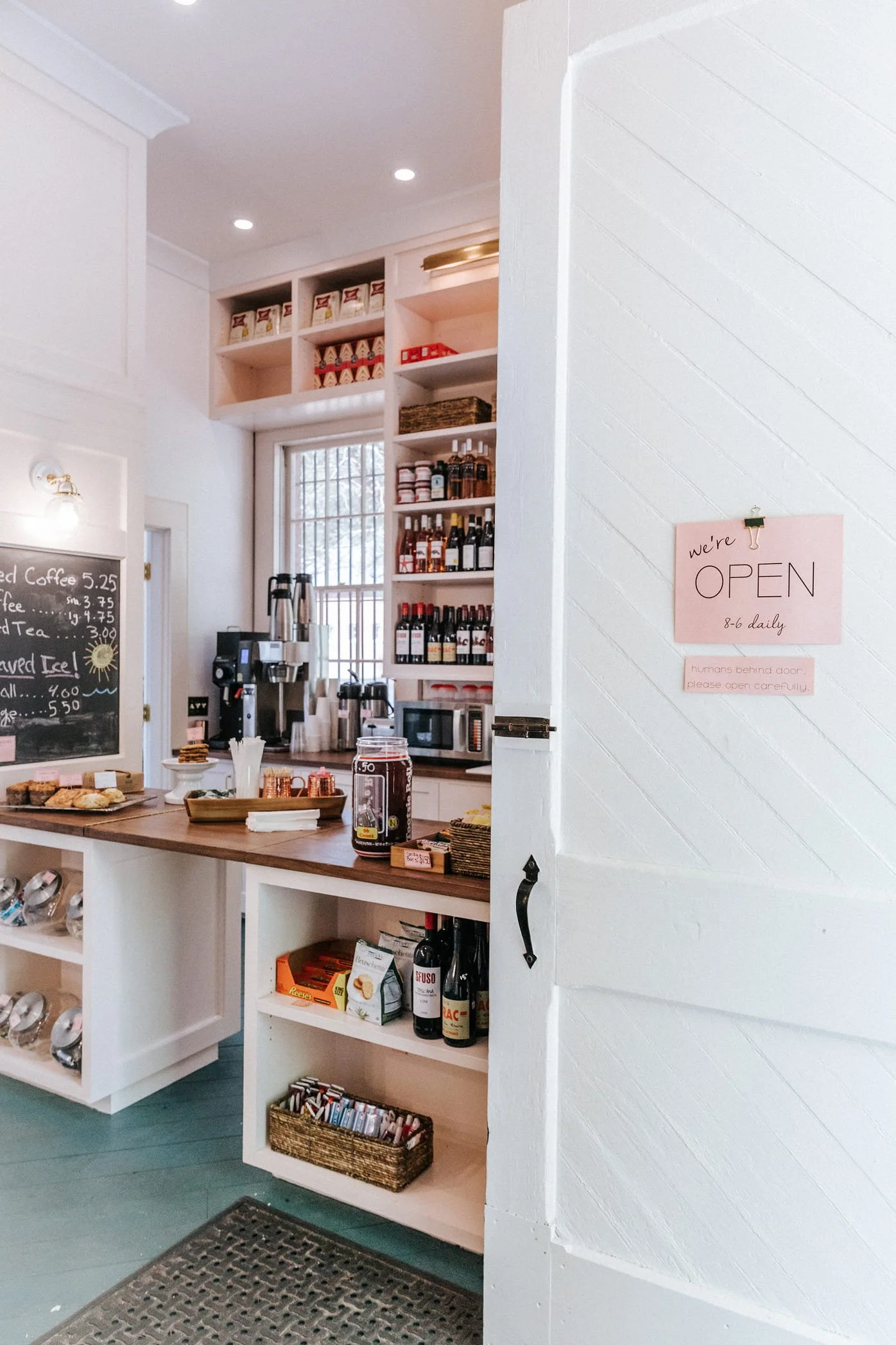 View of a cozy cafe or small restaurant interior with a white wooden counter, shelves stocked with wine bottles, spices, and food items. A sign on the wall reads "we're OPEN 8-6 daily." There are baked goods on the counter, a blackboard menu, and a beverage station with coffee dispensers, a microwave, and cups. The setting appears warm and inviting.