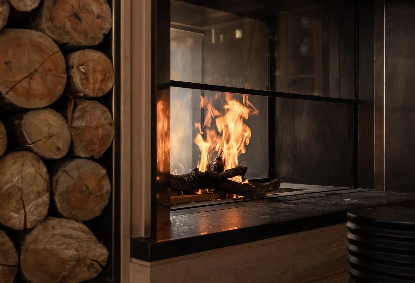 A cozy indoor fireplace with a metal screen and burning logs, adjacent to a neatly stacked pile of firewood.