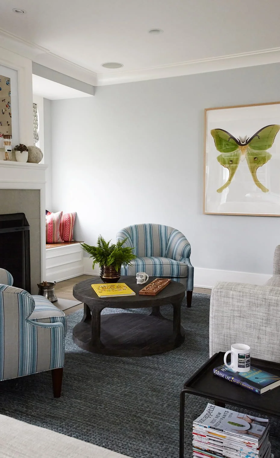 Living room with two striped armchairs, a gray area rug, a round black coffee table with plants and books, a white sofa, and a large framed butterfly print on the wall.