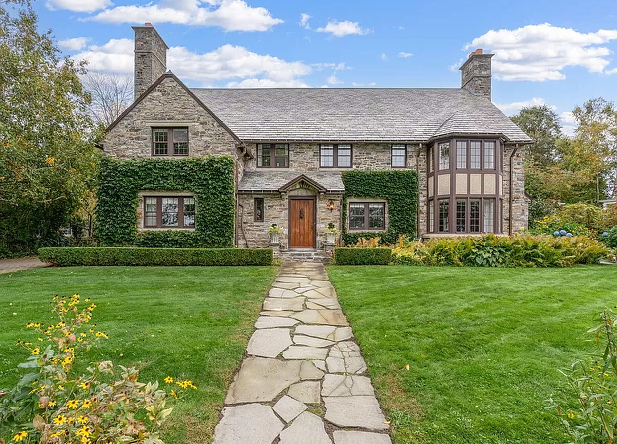 Front view of a large stone house with a lawn and garden, a stone pathway leading to the wooden front door, and trees and bushes surrounding it, under a partly cloudy blue sky.