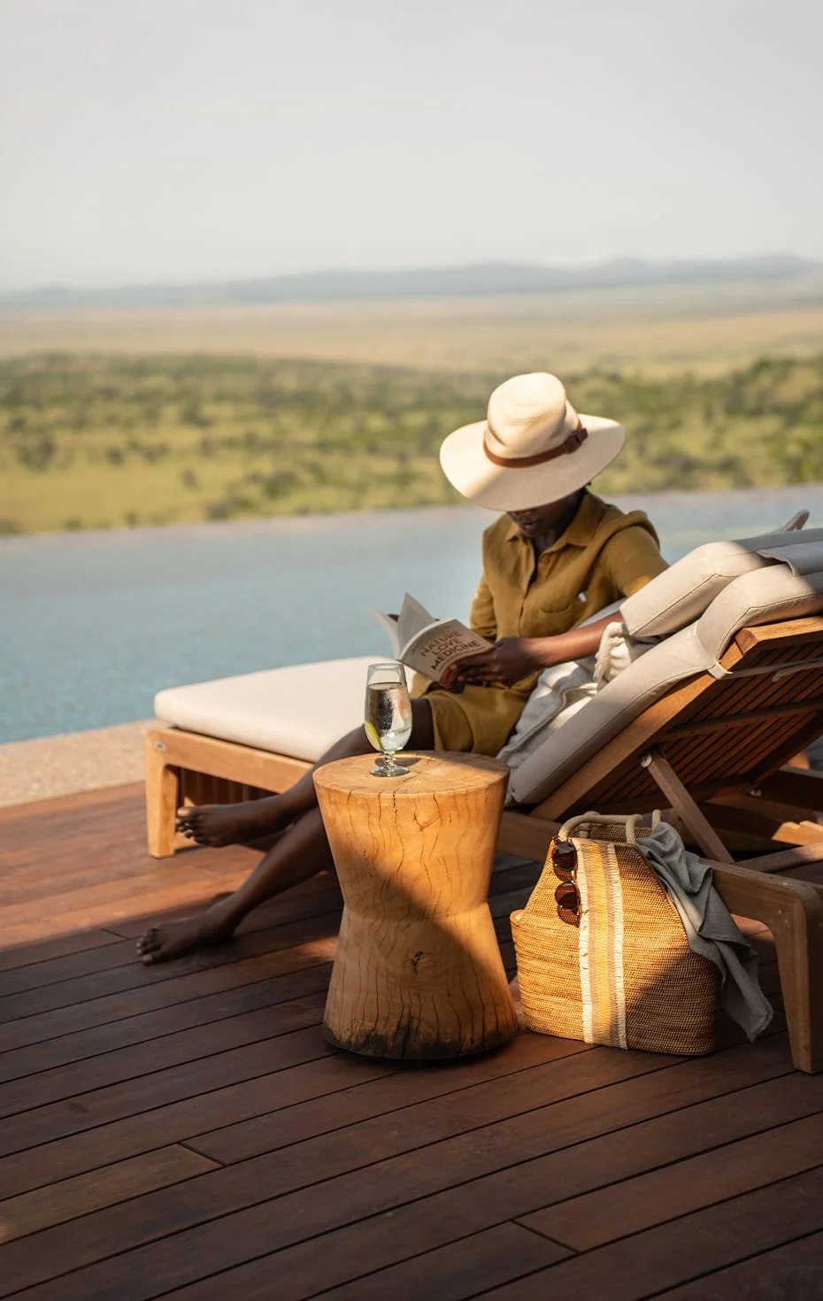 A person relaxing on a lounge chair by a pool, reading a book, wearing a large sun hat and a brown outfit, with a glass of water on a small wooden side table, and a straw bag on the deck.