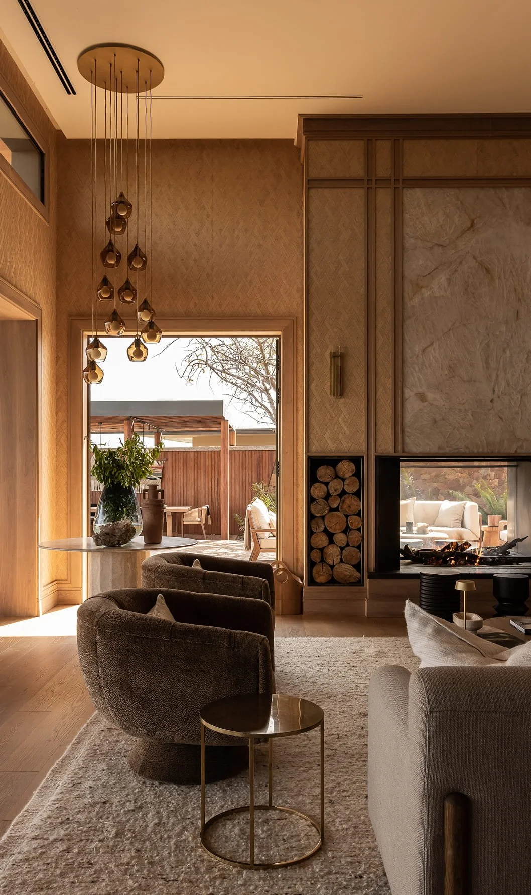 A cozy living room with modern decor, a fireplace, and a glass door leading to an outdoor patio with wooden furniture and a pergola. There are armchairs, a small side table, a large vase with green foliage, and a pendant light fixture hanging from the ceiling.