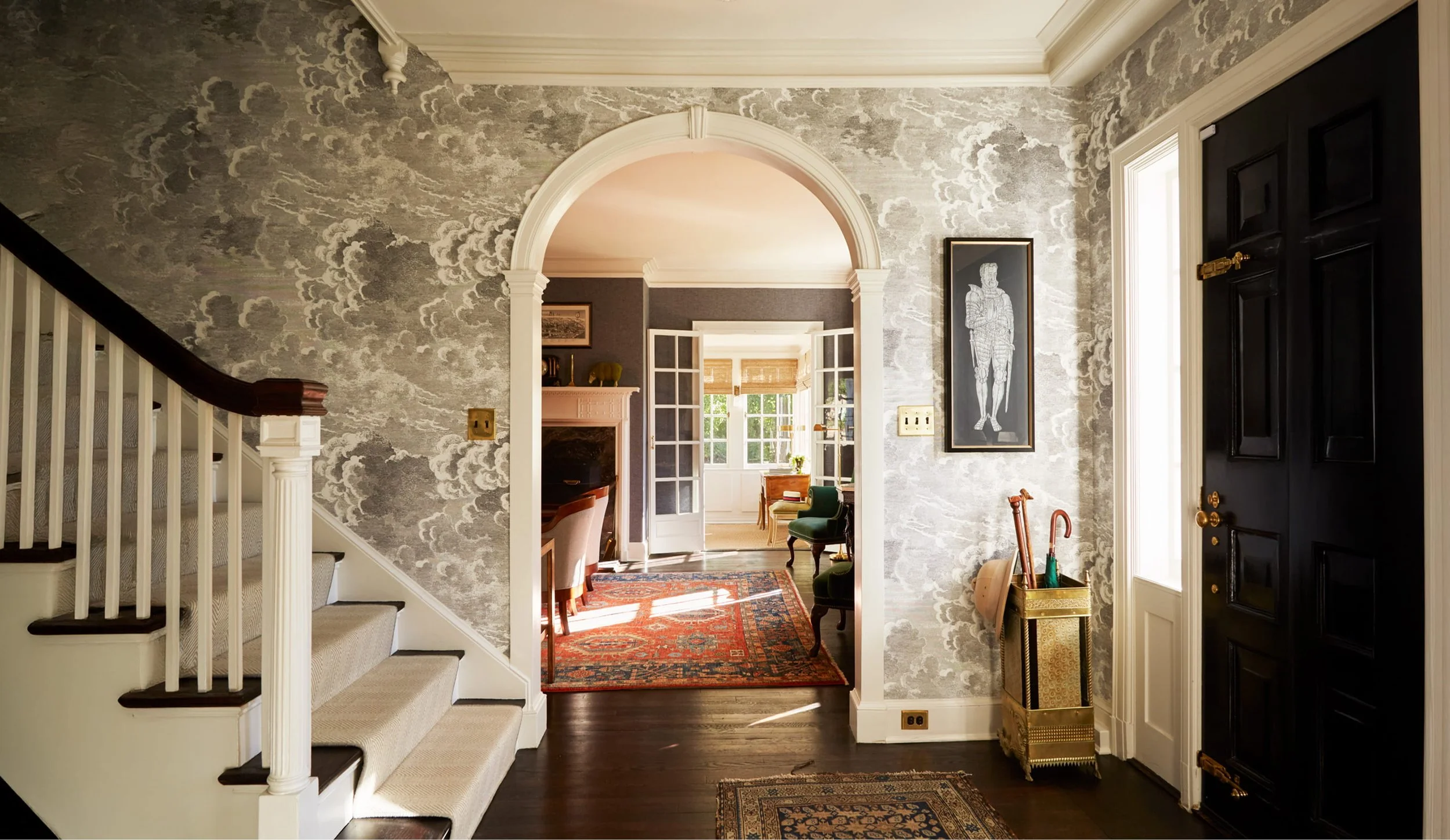 Entrance hall with dark front door, decorative wallpaper, and view into living room with large window, fireplace, and armchairs. Staircase on the left, framed artwork on the wall, and a red patterned area rug.