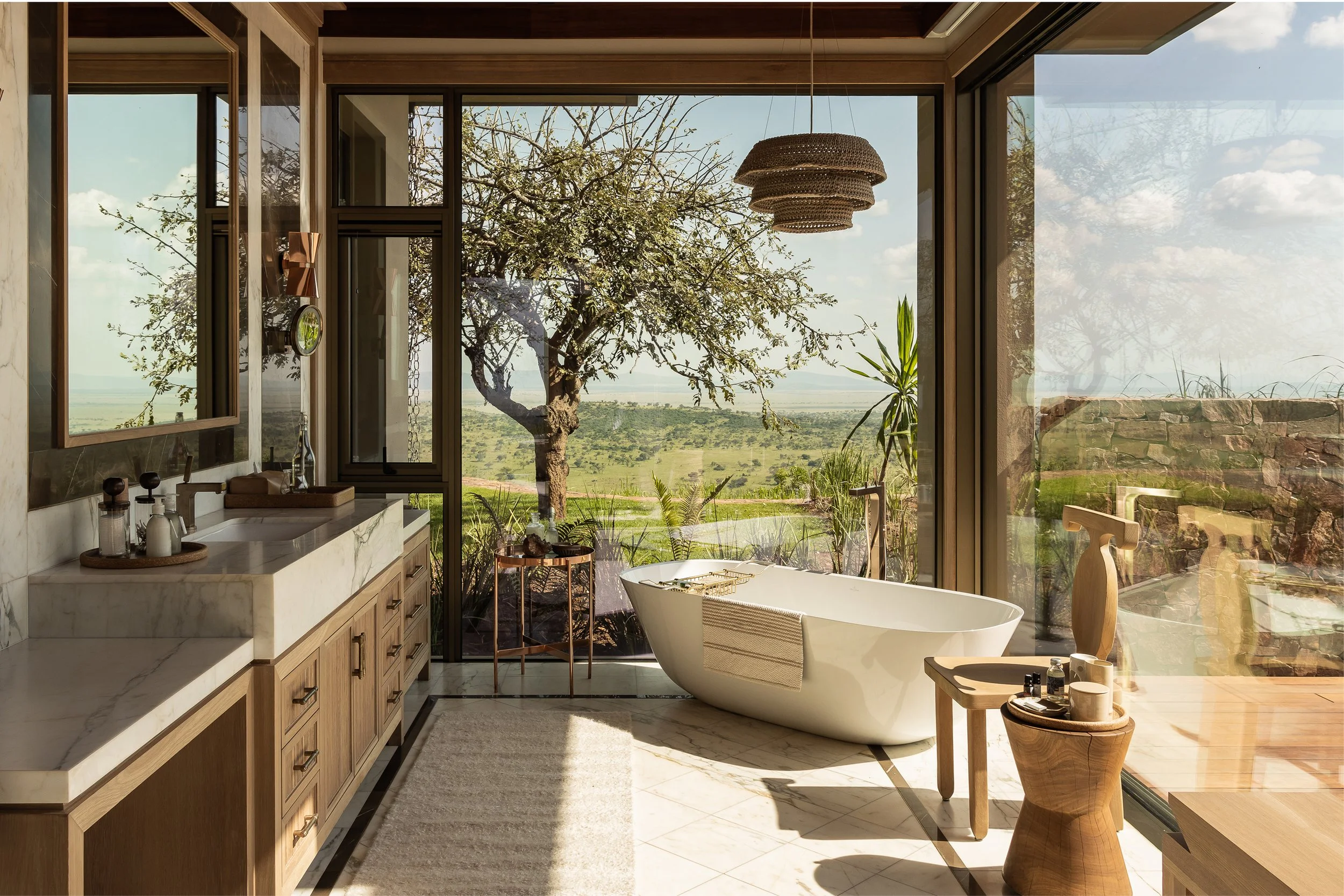 A luxurious bathroom with a freestanding bathtub, large windows showing a landscape with a tree and sky, wooden accents, and modern decor.