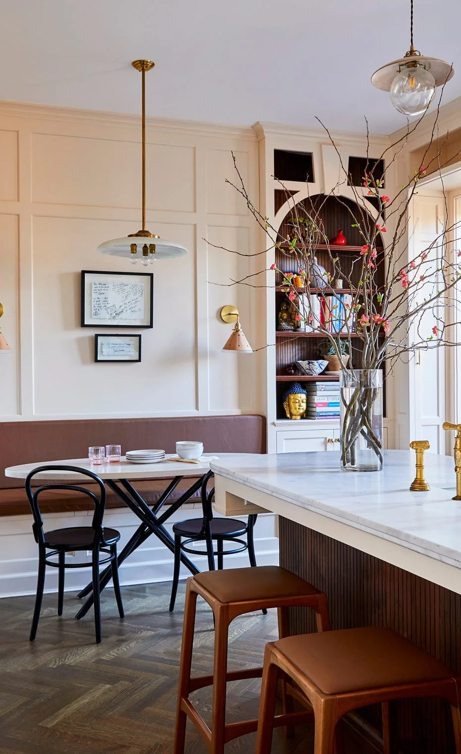 A modern dining room with a white marble kitchen island, black chairs, a built-in bookshelf with decorative objects, and a vase of blooming branches on the island.