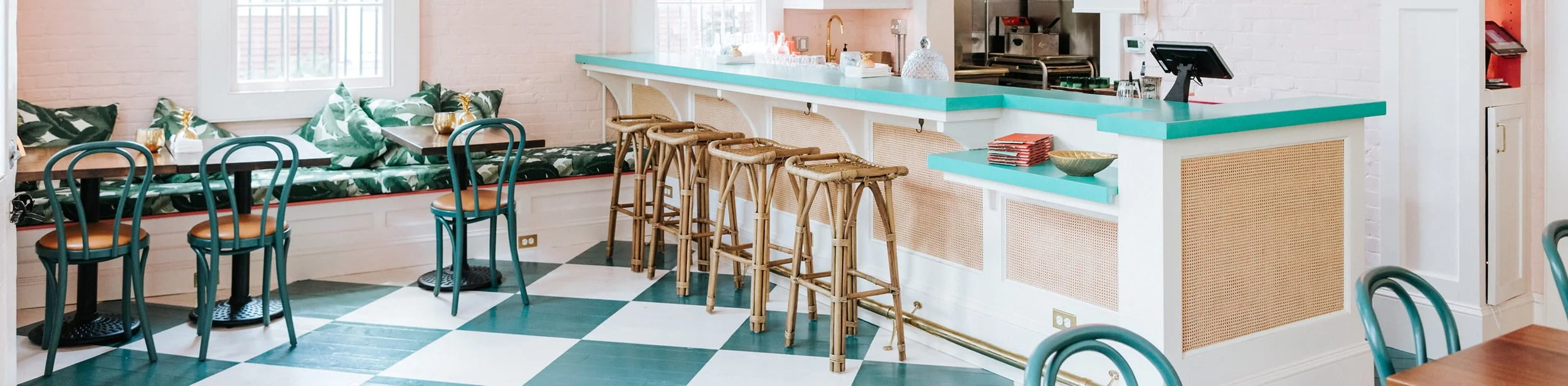 Interior of a cafe with a checkered green and white floor, a white brick wall, and pink accents. There are teal chairs, a built-in bench with tropical leaf cushions, and a counter with a turquoise top and several barstools. The counter has a cash register and some bowls, with cooking equipment in the background.