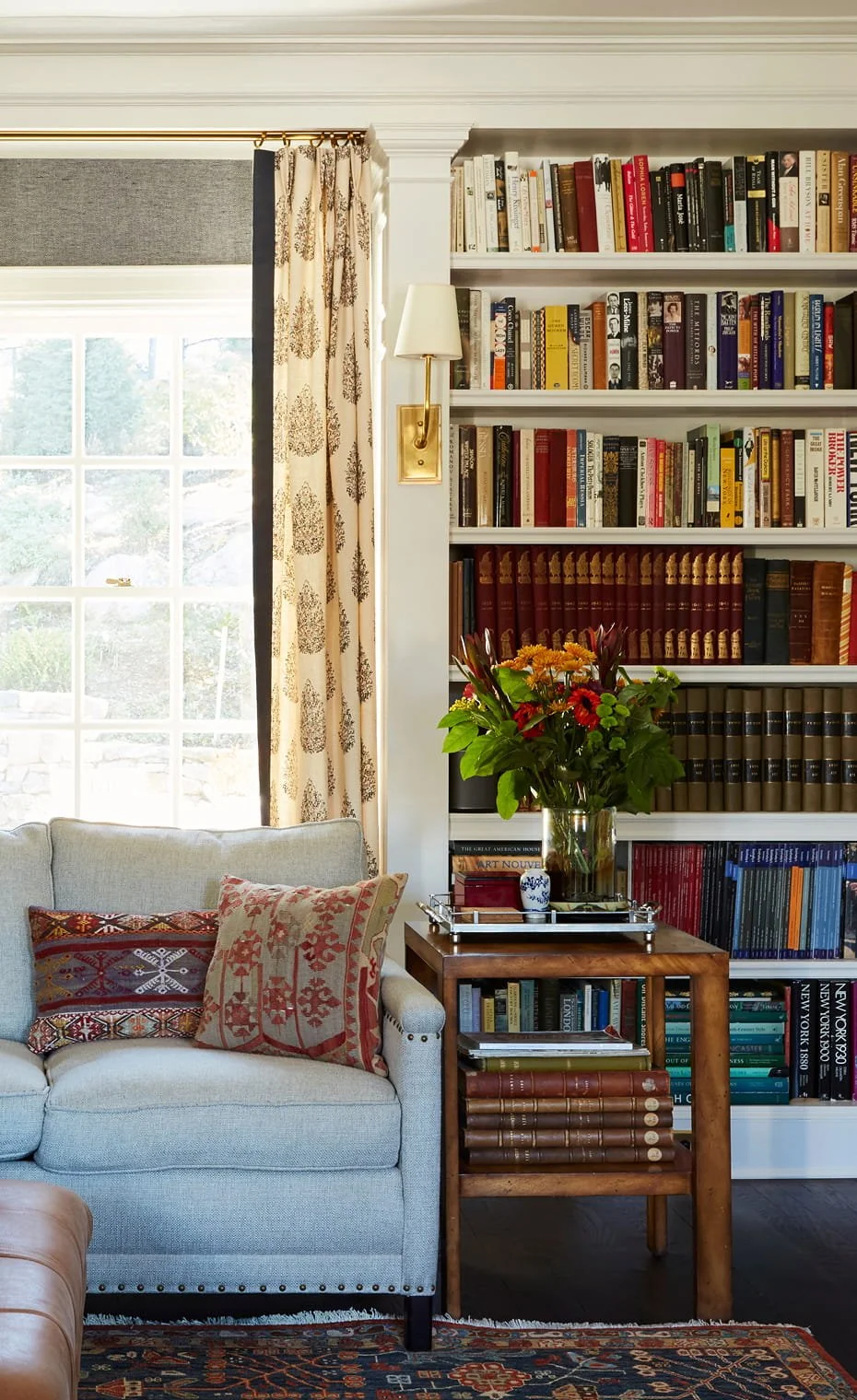 Living room scene with a bookshelf, floral arrangement, sofa with decorative pillows, window with curtains, and a side table with books and flowers.