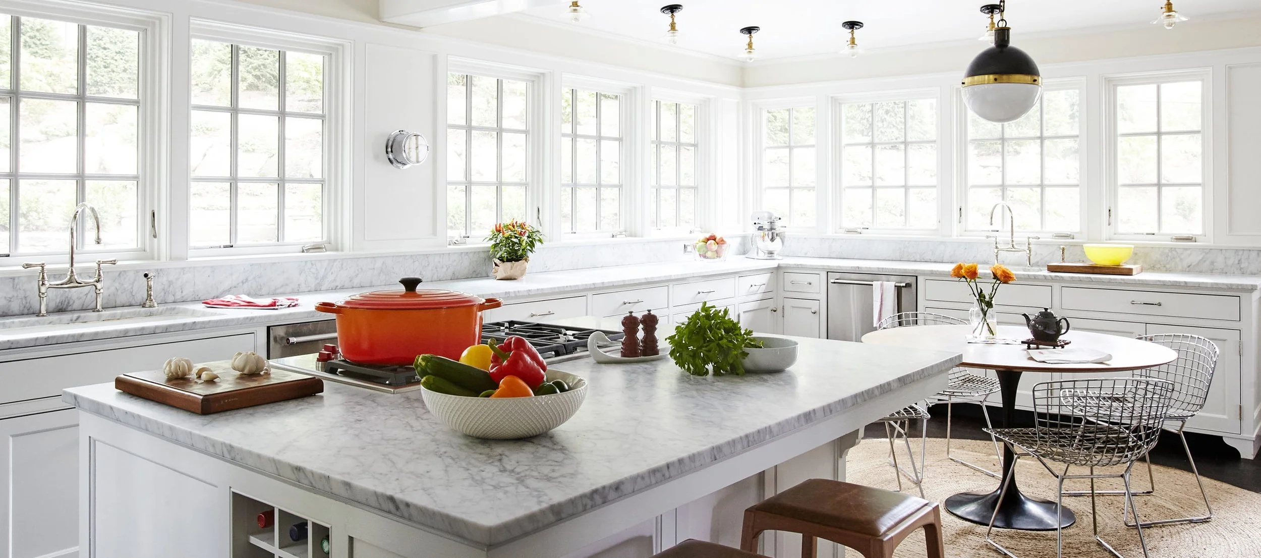 Bright white kitchen with marble countertops and large windows, decorated with fresh vegetables, flowers, and kitchen appliances.