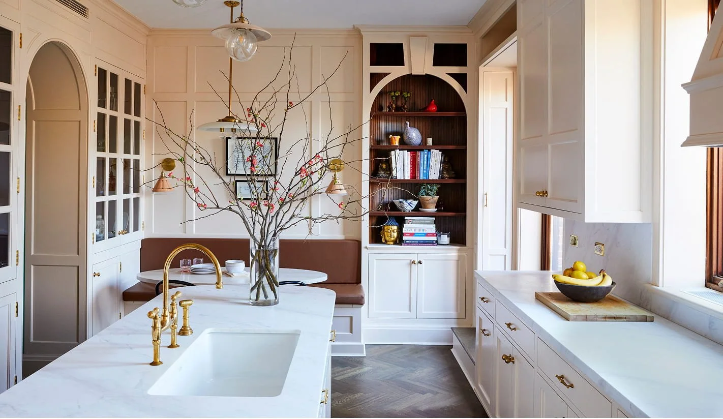 Bright kitchen with white cabinets, marble countertops, and a large island. A glass vase with bare tree branches and pink blossoms is on the island. Open shelving displays books, decorative items, and pottery. A bowl of bananas and apples is on the windowsill, with natural light illuminating the space.