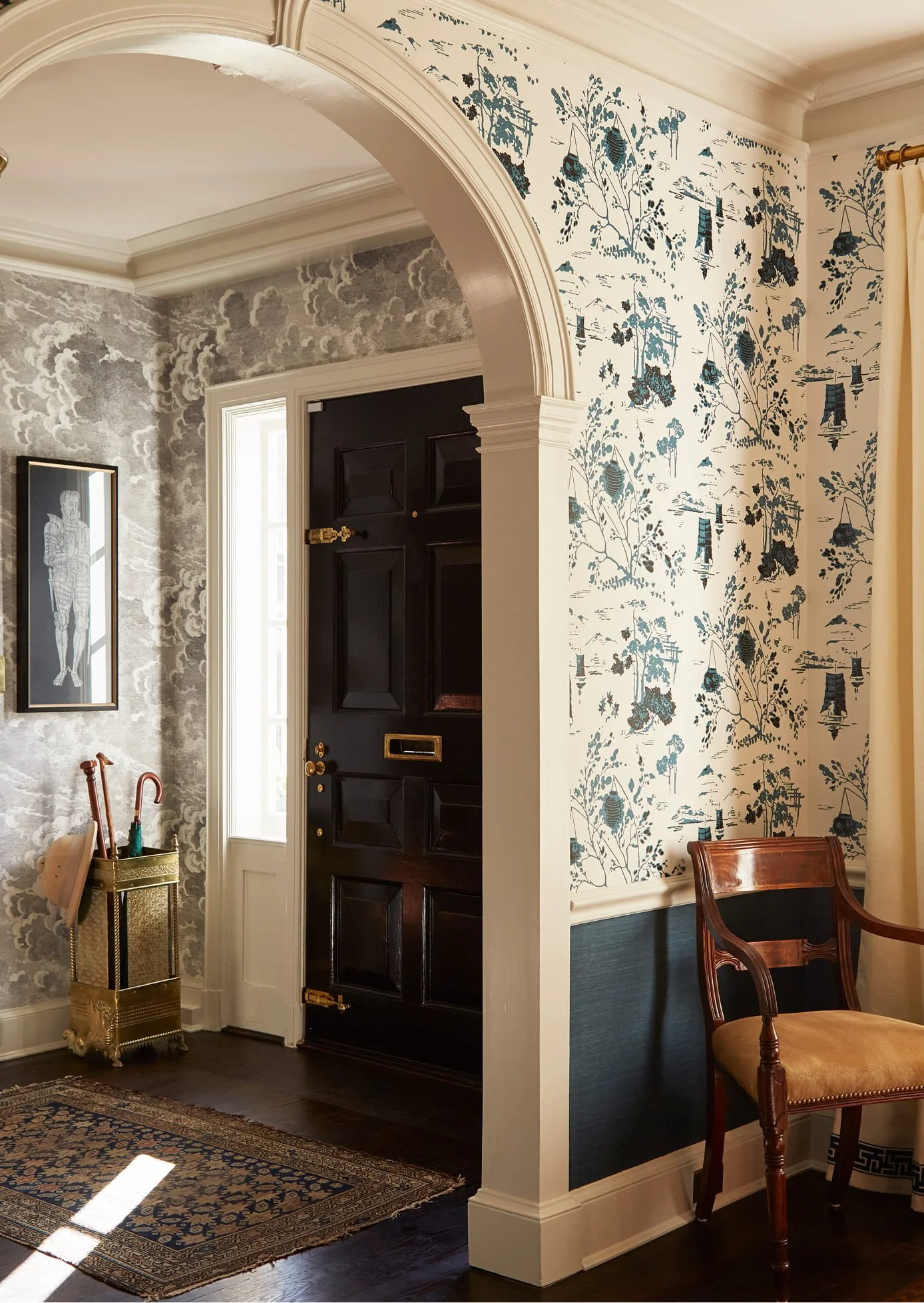 Interior view of a room with an arched doorway, dark wood front door, and patterned wallpaper. There is a small table holding umbrellas and a framed picture on the wall. A chair with wood frame and upholstered seat is partially visible.