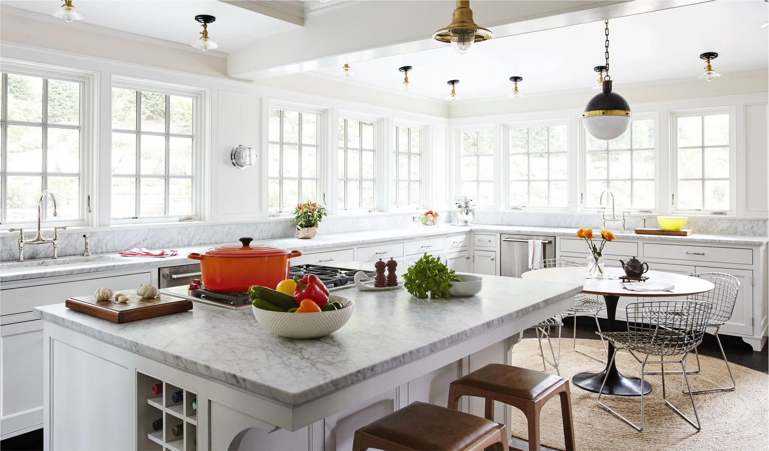 Bright kitchen with white cabinetry and marble countertops, featuring a large central island with a red pot, vegetables, and herbs, surrounded by chairs, and multiple windows letting in natural light.