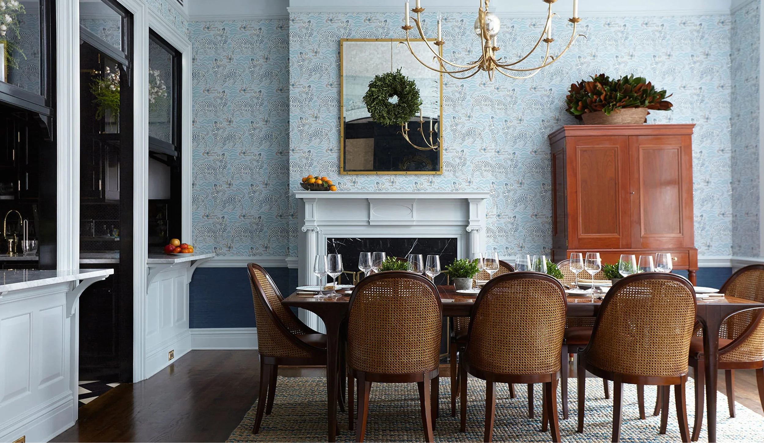 A dining room with a wooden table set with wine glasses and small potted plants. There are rattan-back chairs around the table. The room has light blue patterned wallpaper, a gold-framed mirror, a white marble fireplace, and a wooden cabinet with a plant on top. A chandelier hangs from the ceiling.