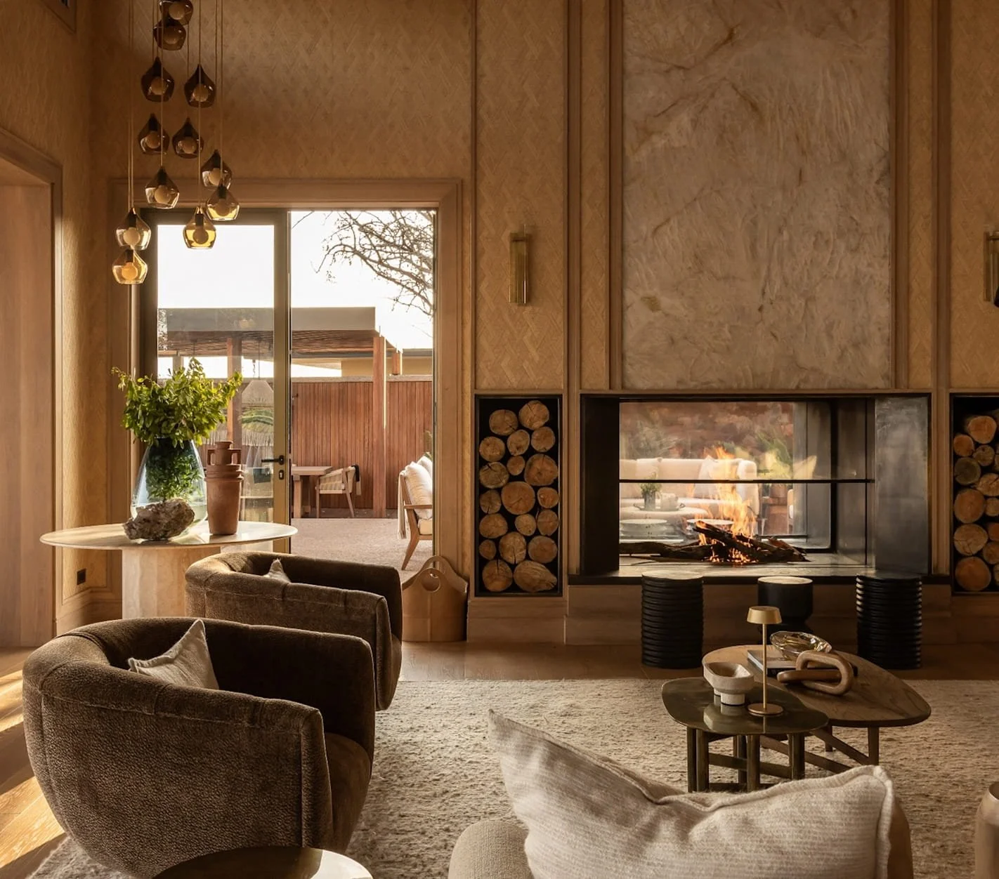 Living room with a stone fireplace, wood logs on either side, a fire burning inside, a round coffee table with decorative items, upholstered chairs, a large plant and vases on a side table, and a patio visible through a glass door with outdoor furniture.