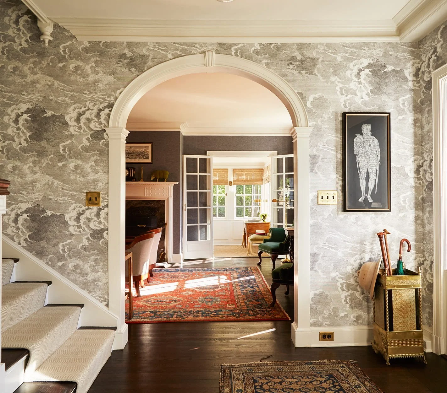 Interior view of a house with a staircase on the left, a decorative wallpapered wall, an arched doorway, and a room in the background with chairs, windows, and a fireplace.