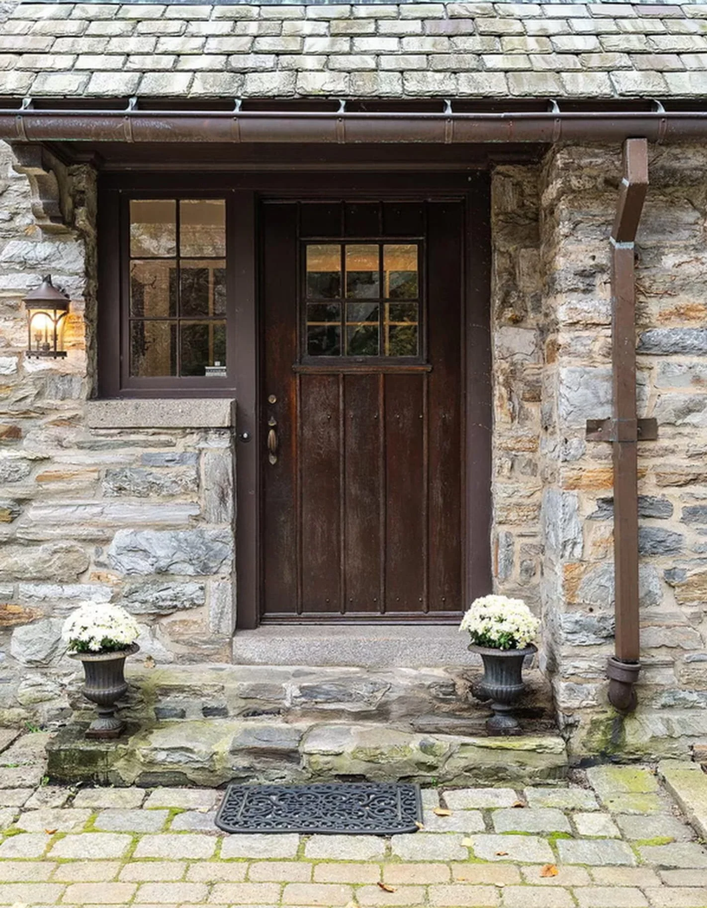 Front door of a house with a stone exterior, a small window on the door, two potted white flowers on either side of the door, a porch light to the left, and a doormat in front.