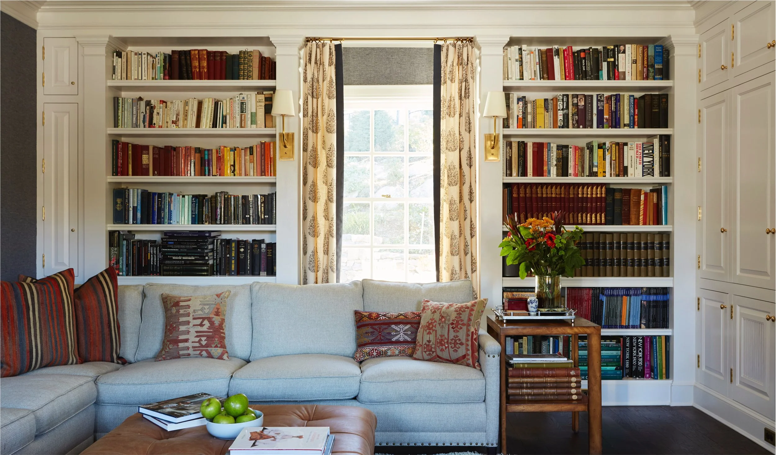 Living room with white bookshelf filled with books, beige curtains, and a large window. A beige sofa with throw pillows in front of the bookshelf. A wooden coffee table with green apples, books, and magazines. A glass vase with flowers on a side tabl