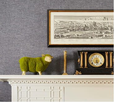 Close-up of a white mantle with decorative molding, featuring a green plush sheep, a tall gold candlestick, a small black vase, a black and gold clock, and a framed cityscape drawing on a gray wall.