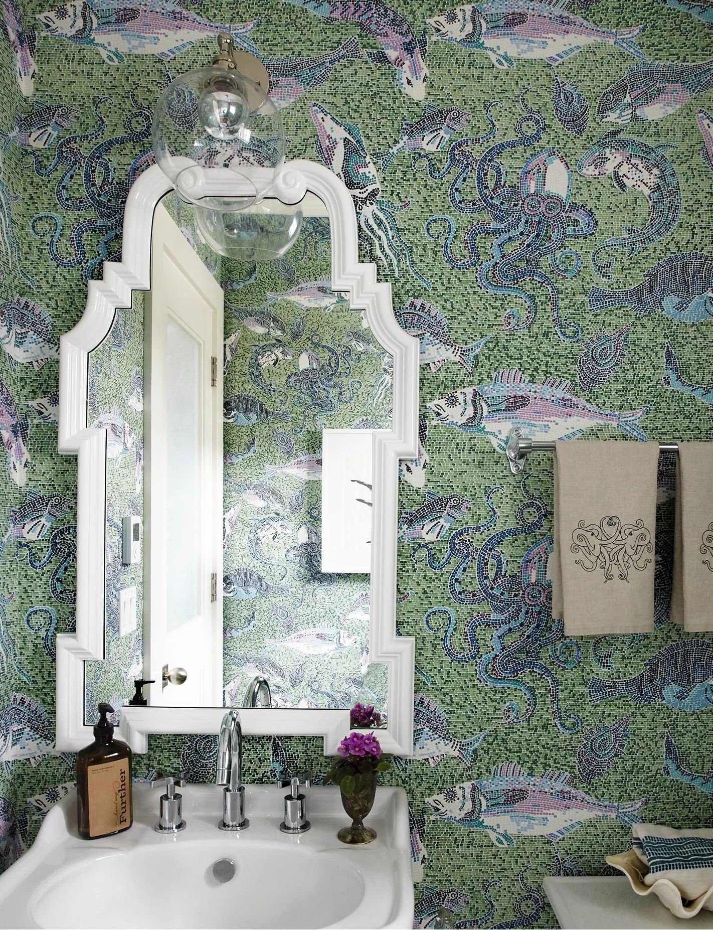 Bathroom with mosaic wall featuring fish, sharks, and an octopus, white framed mirror, sink with silver faucet, purple flowers in a copper pot, brown soap dispenser, towel rack with beige and blue towels.