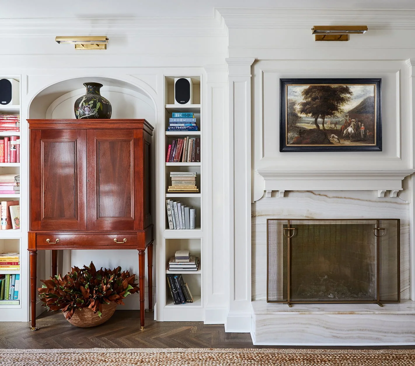 Living room with white built-in bookshelves, a wooden cabinet, a framed landscape painting above a fireplace, and a potted plant on the floor.
