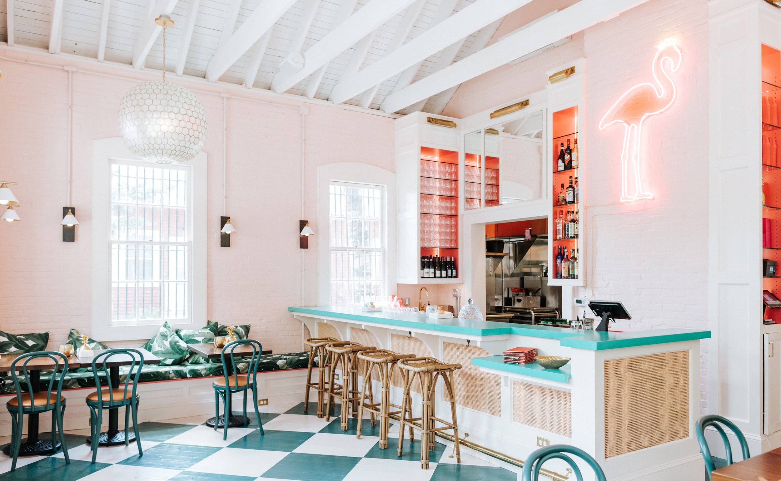 Interior of a bright, modern cafe with white brick walls, large windows, and a high ceiling with exposed beams. There is a bar area with barstools, tropical-patterned couches, and various tables and chairs. A neon flamingo sign is on the wall above the bar.
