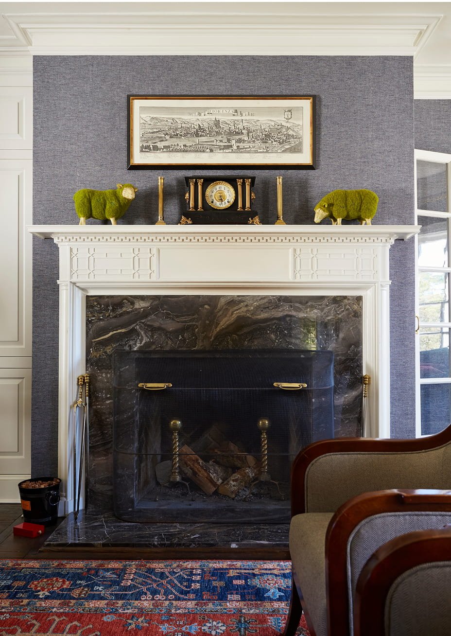 Decorative living room fireplace with white mantel and black marble surround, flanked by brass andirons, with a dark gray textured wall above. The mantel holds two green moss sheep, two tall gold candles, a black antique clock, and a framed black-and-white cityscape drawing. Part of a wooden chair and a colorful patterned rug are visible in the room.