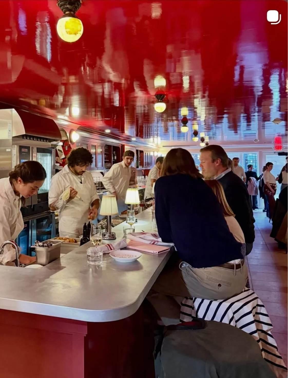A lively restaurant bar with patrons and chefs, featuring a red ceiling and warm lighting.