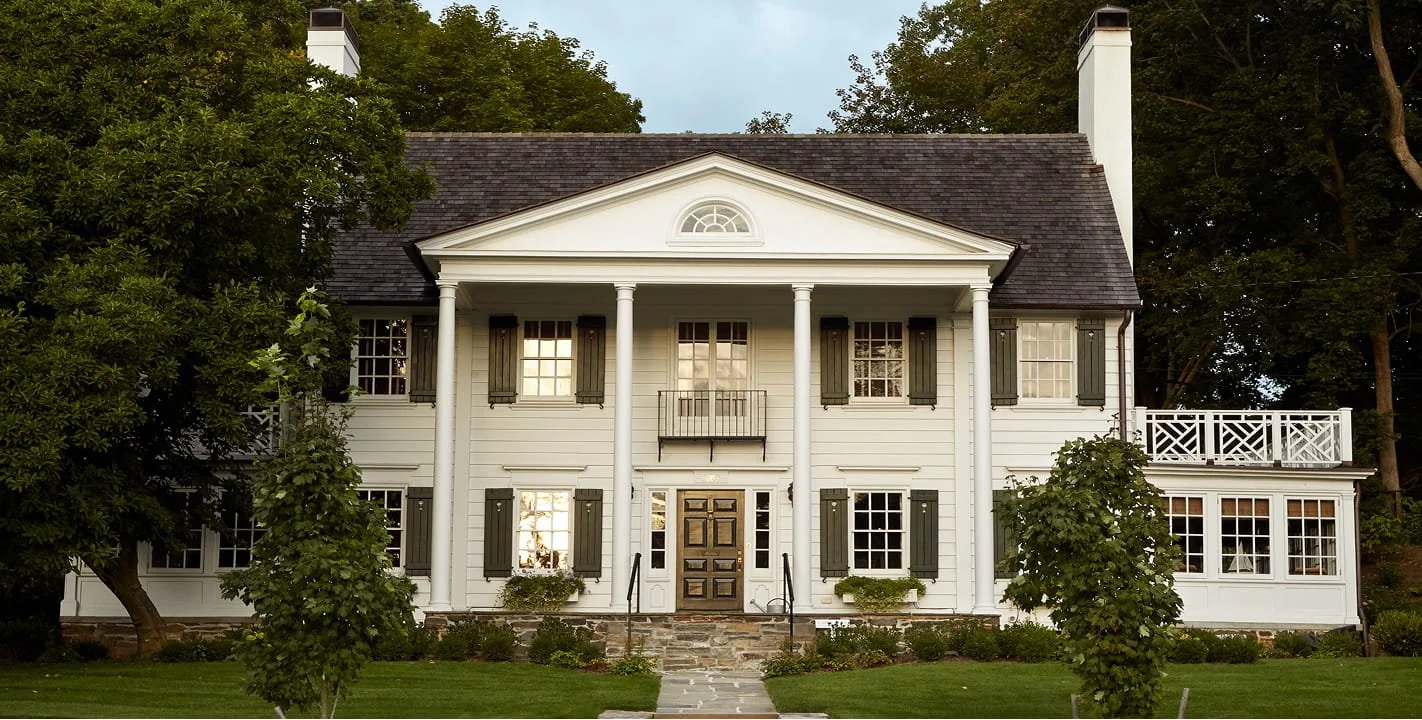 Large white colonial-style house with black shutters, columns, and a balcony. Surrounded by trees and a well-manicured lawn.