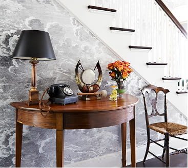 A wooden console table with a black lamp, a rotary phone, a decorative mirror, a flower arrangement, and a brass decorative object, placed in front of a gray textured wall and beside a staircase with white risers and dark wood treads.