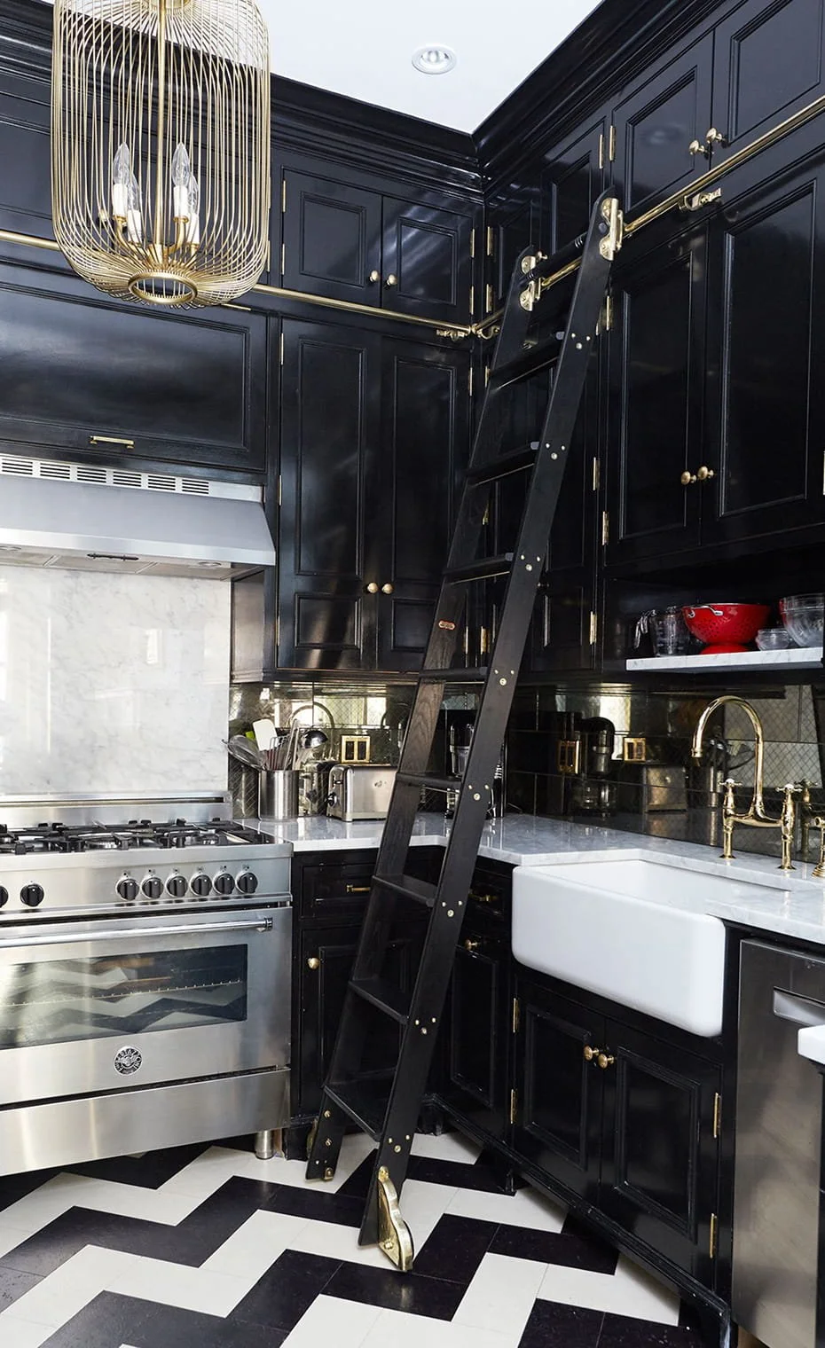 A modern kitchen with black cabinets, a white farmhouse sink, gold hardware, a stainless steel stove, a black ladder leaning against the cabinets, black and white chevron-patterned floor, and a gold chandelier.