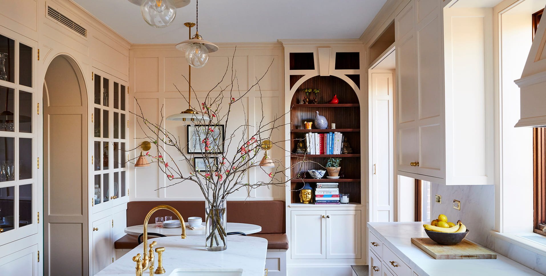 A bright, elegant kitchen and dining area with white cabinets, a marble countertop, and a round dining table with a vase of branches and pink flowers, surrounded by various decor and books.