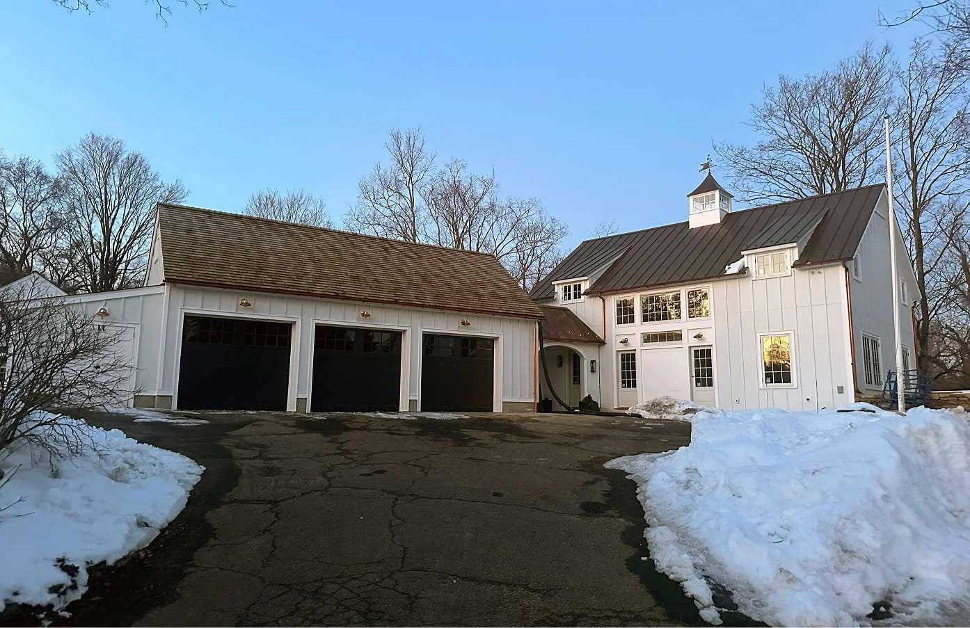 A large white house with a garage and snow on the ground. The house has multiple windows, a metal roof, and a small cupola with a weather vane. There's snow on the driveway and surrounding areas, with leafless trees in the background.