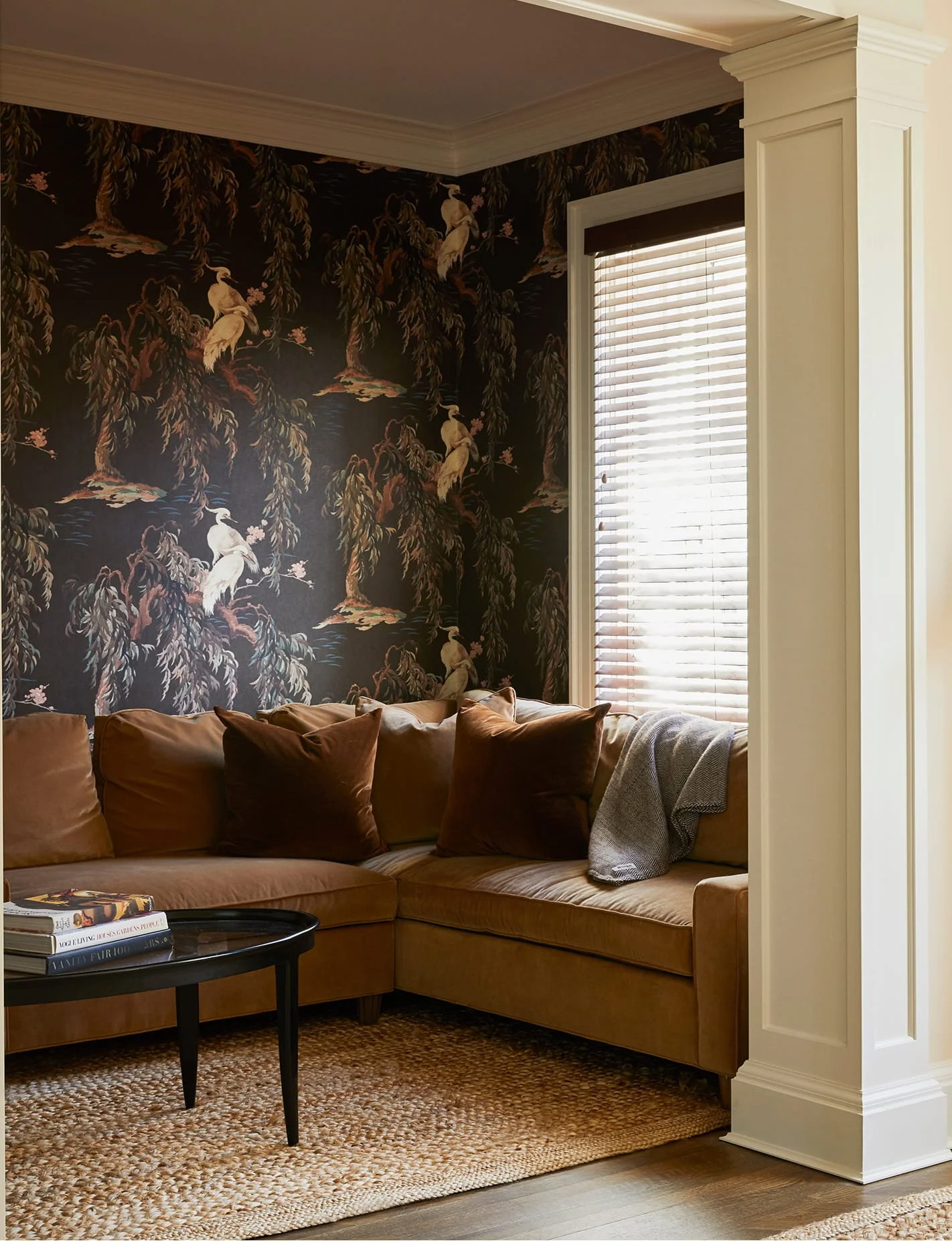 A living room corner with a beige sectional sofa with brown and beige pillows, a woven rug, a black coffee table with books, a window with blinds, and a wallpapered wall with a nature scene of trees and birds.