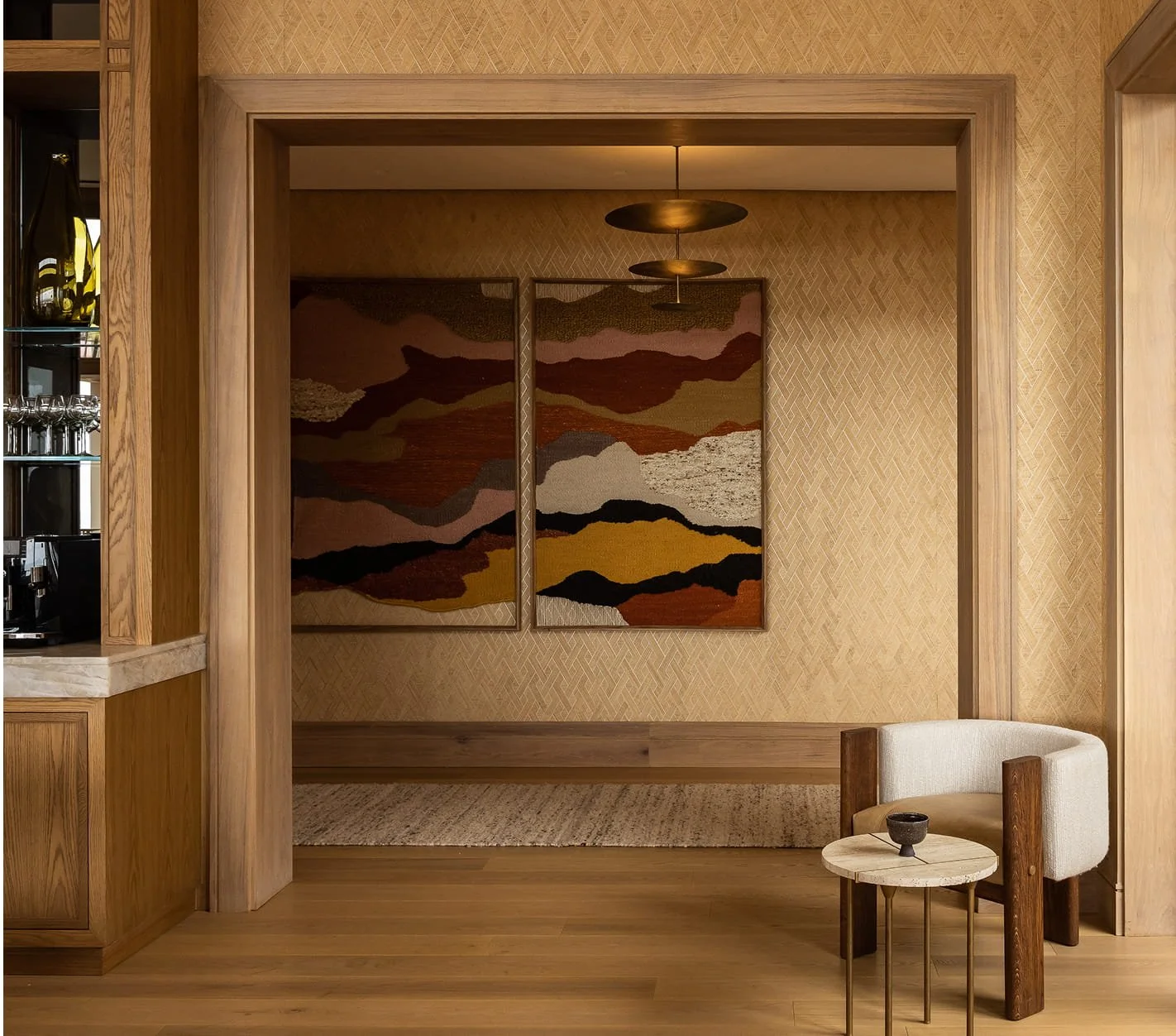 A cozy interior space with wooden walls, a woven rug, and abstract landscape artwork. There is a small beige and brown armchair next to a round side table with a dark bowl. Part of a bar or kitchen area is visible on the left.