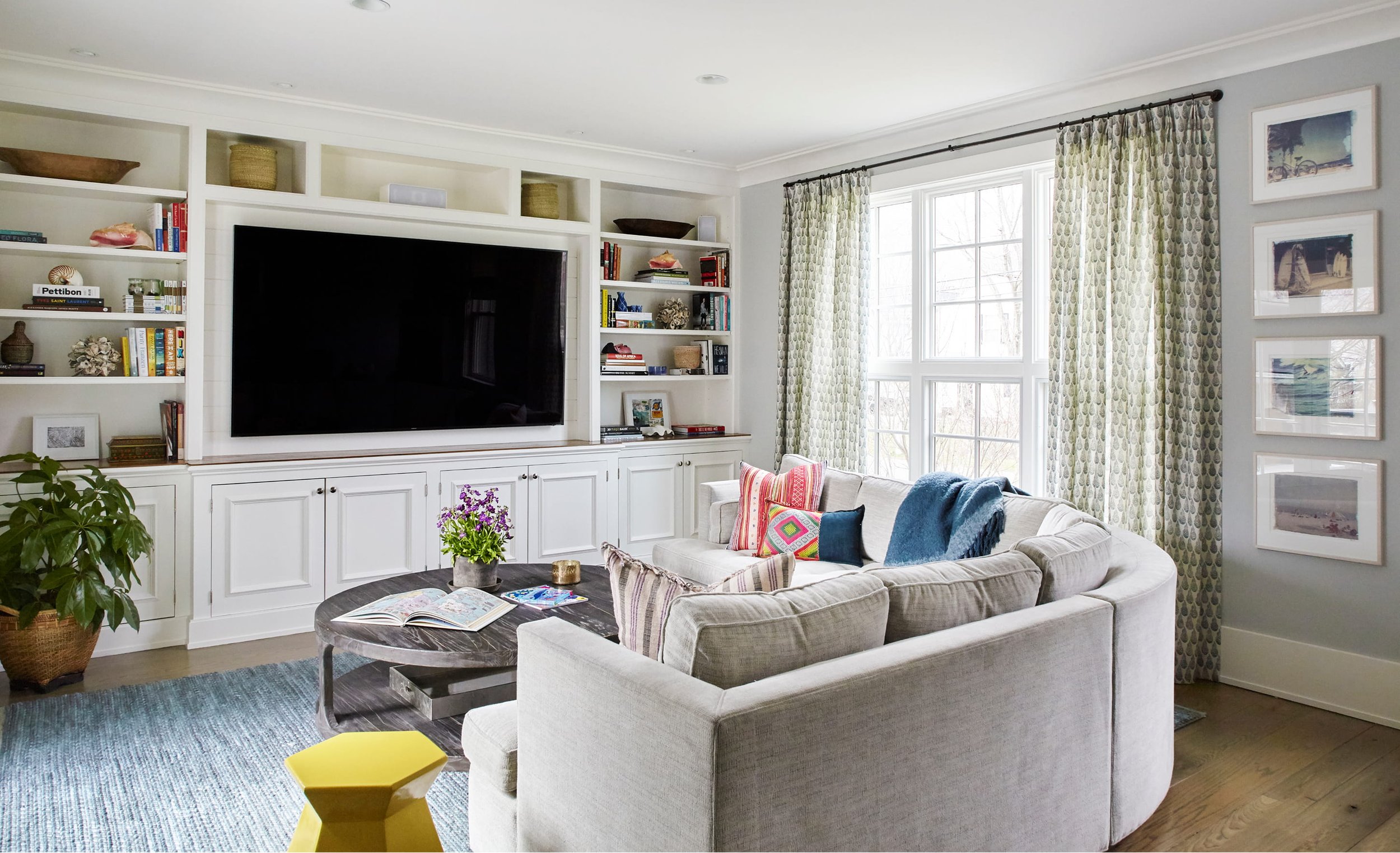 A bright, cozy living room with built-in white shelving filled with books and decorative items, a flat-screen TV, and a large window with patterned curtains. There is a white sofa with colorful pillows, a round wooden coffee table with a flowerpot and magazines, and a blue rug on a wooden floor.