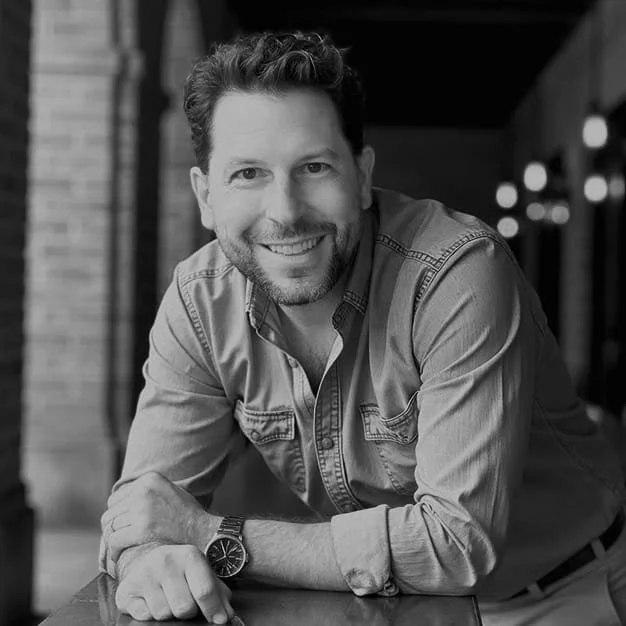 A smiling man with short hair and a beard, wearing a button-up shirt, leaning on a table in a dimly lit indoor setting