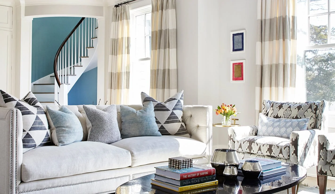 Living room with white sofa, patterned pillows, beige and gray armchair, coffee table with books and decorative objects, window with striped curtains, and staircase with teal wall in the background.
