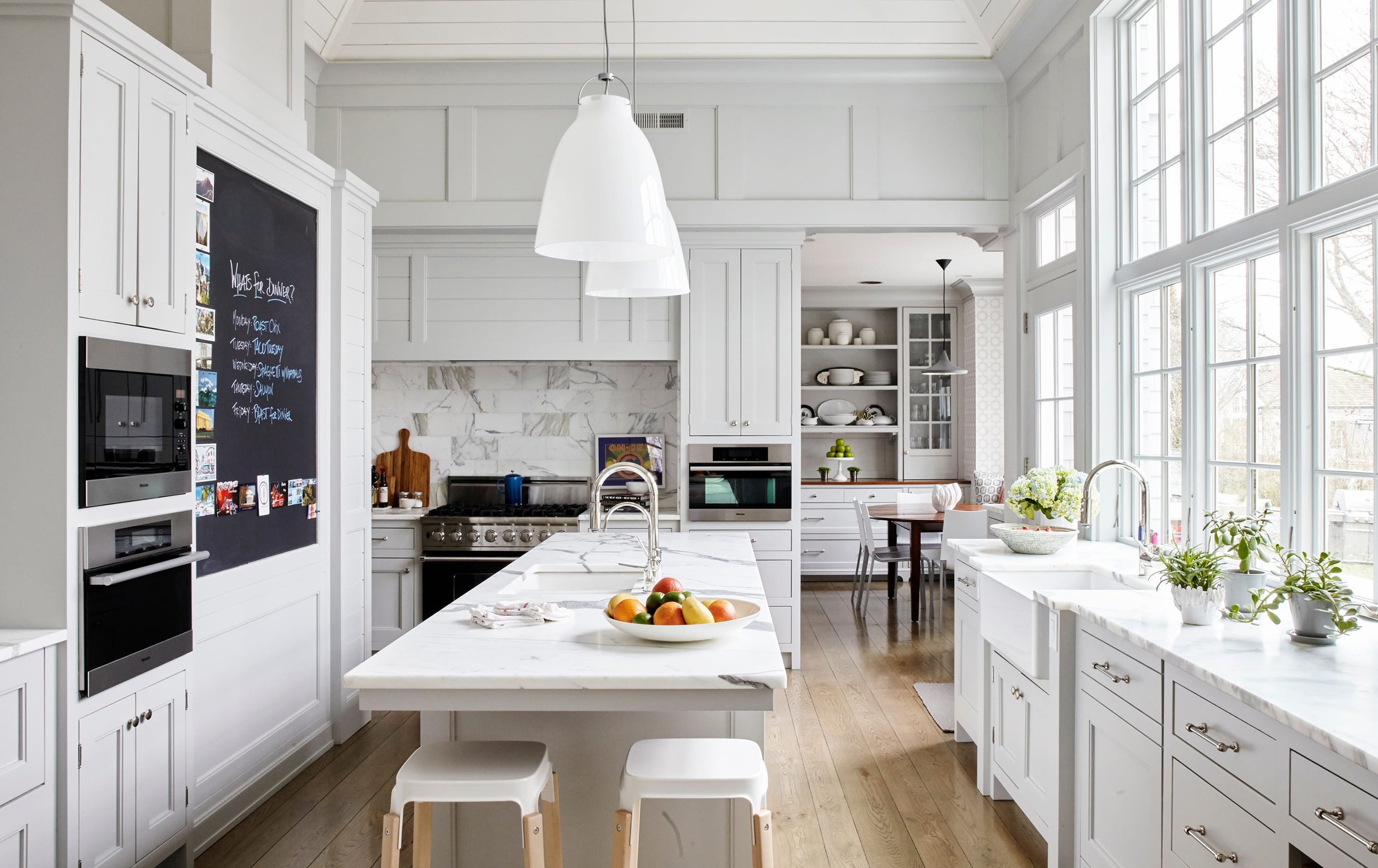 Bright, modern white kitchen with large windows, marble countertops, and wooden flooring. Contains a kitchen island with a bowl of assorted fruits, and a dining table with chairs. Features built-in appliances and open shelving with dishes and decor. Pendant lights hang from the ceiling.