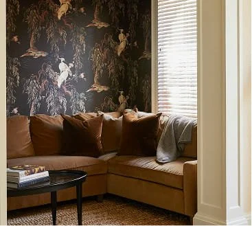 A beige sectional sofa with brown pillows and a gray throw blanket in a living room, next to a window with blinds. There is a black coffee table with books in front of the sofa and dark patterned wallpaper in the background.