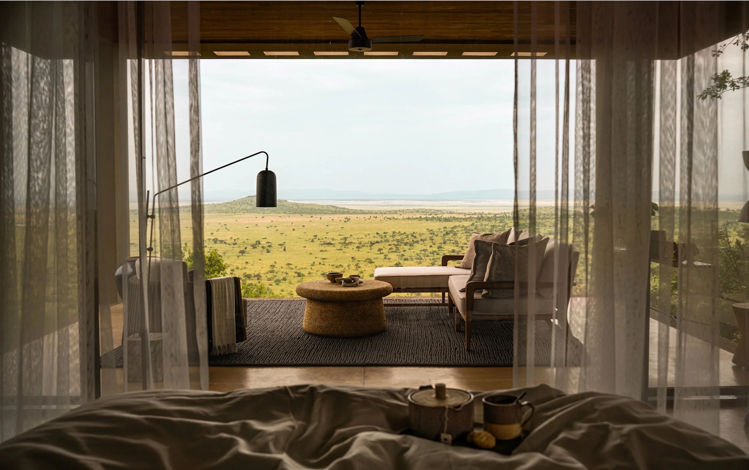 A view from a bedroom looking out onto a living space with an open wall, revealing a vast grassy landscape with sparse trees and distant hills under a cloudy sky.