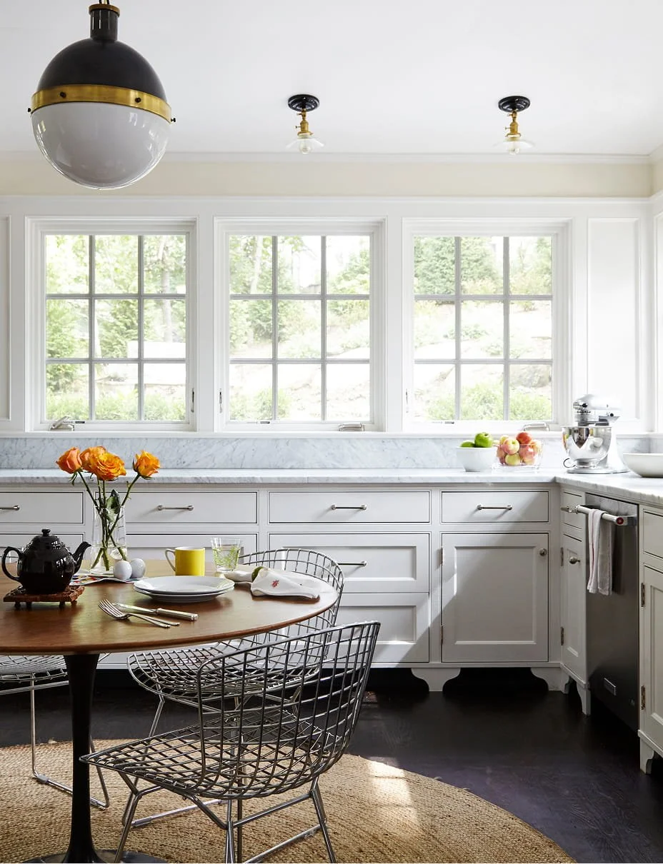 Bright kitchen with three large windows, white cabinets, and dark hardwood floors. A round wooden table with wire chairs, set for breakfast with plates, cutlery, a teapot, and a flower vase. Sunlight streams through the windows, and there are bowls of fruit and a stand mixer on the countertop.