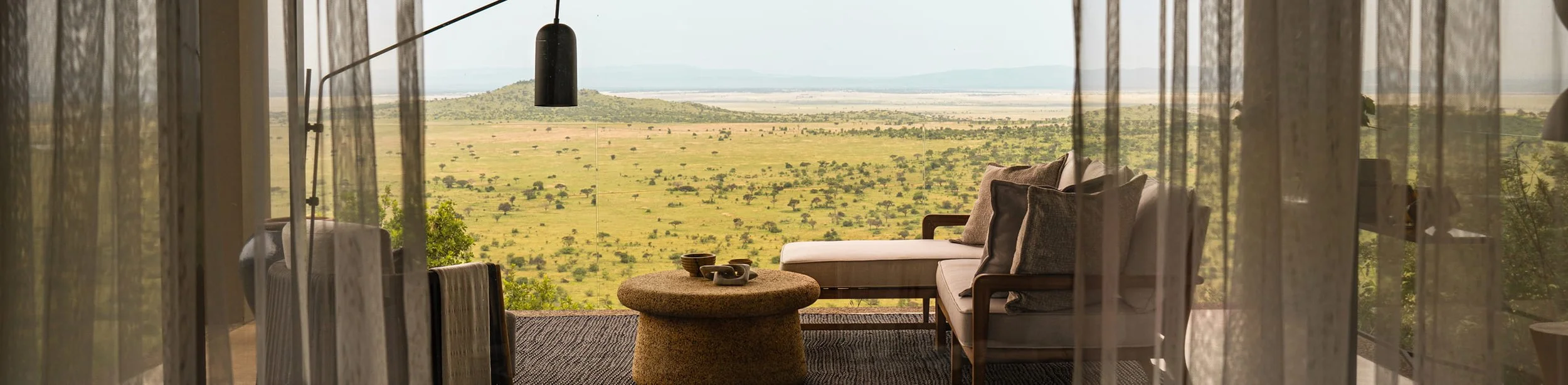 Living room with large windows overlooking an expansive savannah landscape with scattered trees and hills in the distance.