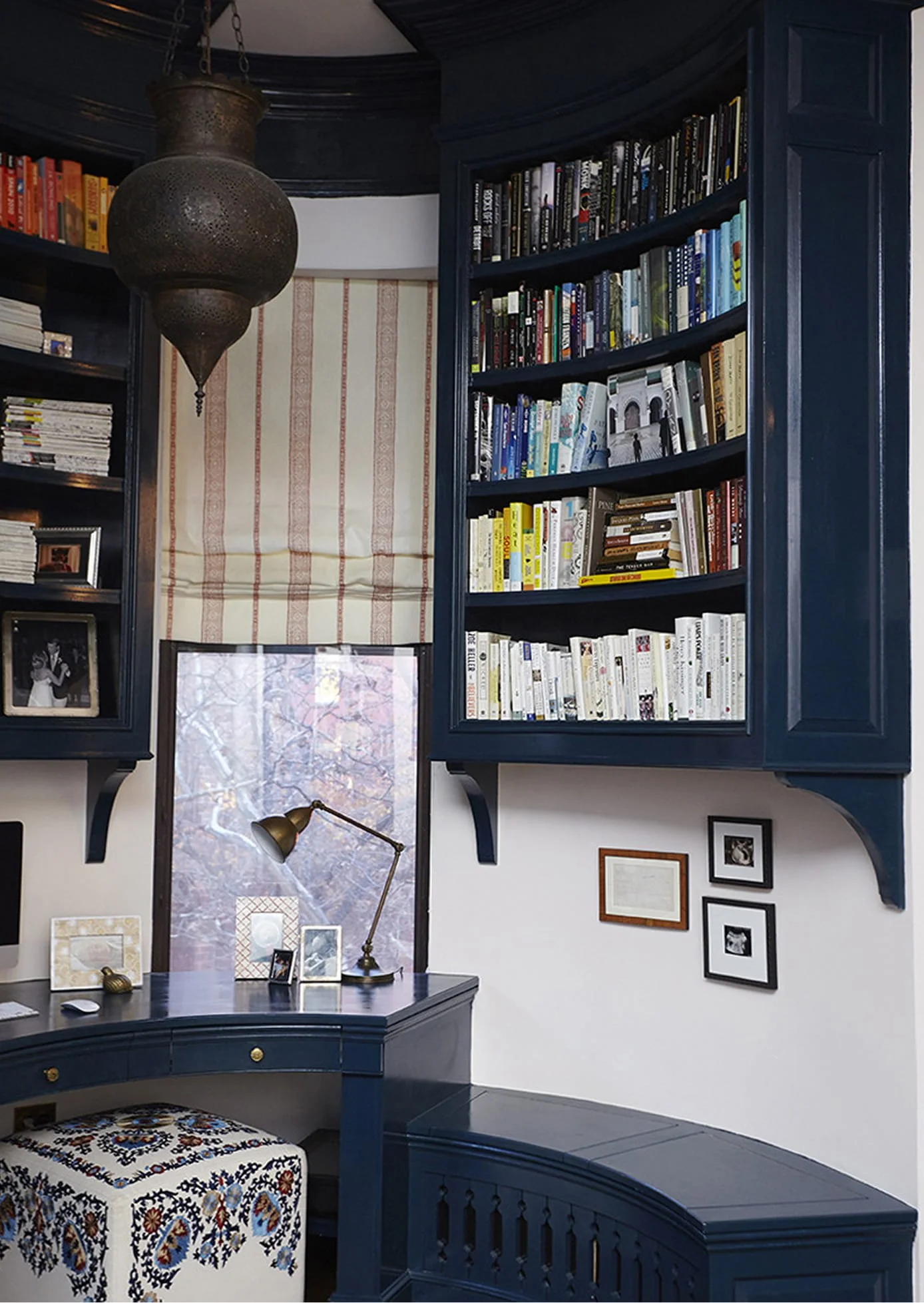 View of a room corner with two black bookshelves filled with books, a window with a beige curtain, a brass desk lamp, and a framed black-and-white photograph on a white wall with several smaller black-and-white framed pictures.