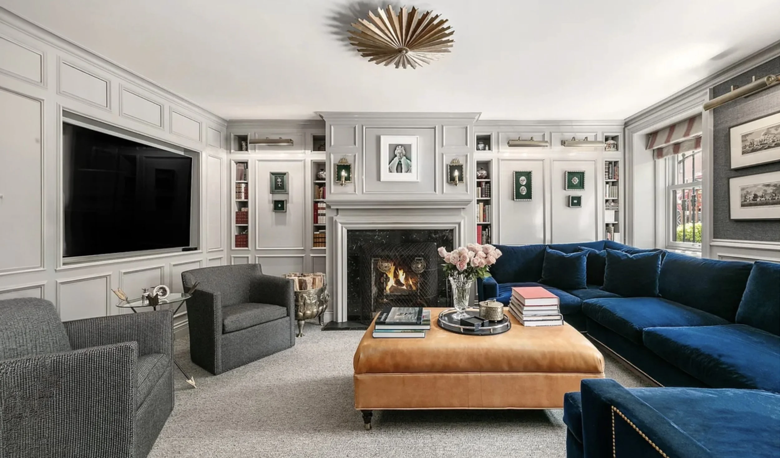 Living room with white paneled walls, built-in shelves, and a decorated fireplace mantel. There is a navy blue sectional sofa, a tan ottoman, and a small round table with books and decorative items. A black armchair and a glass-top side table are als