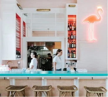 Interior of a modern cafe with a turquoise bar counter, wicker bar stools, and a white wall with a neon flamingo sign; two women are working behind the counter.