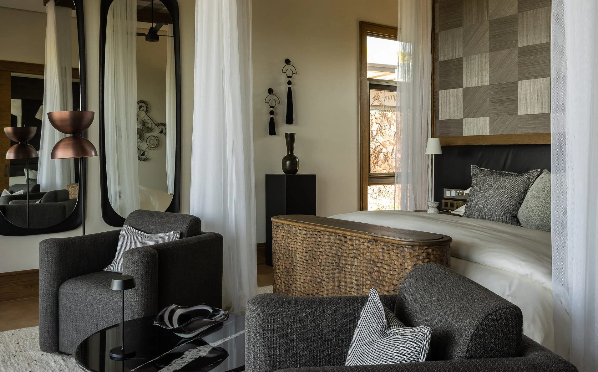 A living room with a bed, gray armchairs, a large mirror, and various wall decorations including a black vase on a black side table and three wall hangings. There are white curtains, a window with sunlight, and a textured wall panel behind the bed.