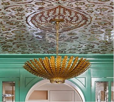 Gold chandelier hanging from an ornate ceiling with floral patterns, above a mint green archway in a decorated room.