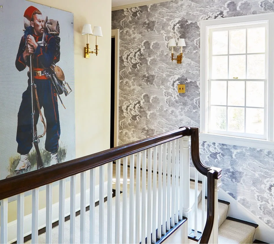 A staircase landing with a large window letting in natural light, decorated with textured gray cloud wallpaper, and featuring a vintage-style wall sconce on a cream wall. A painting of a young boy dressed as a soldier leaning on a rifle is visible.