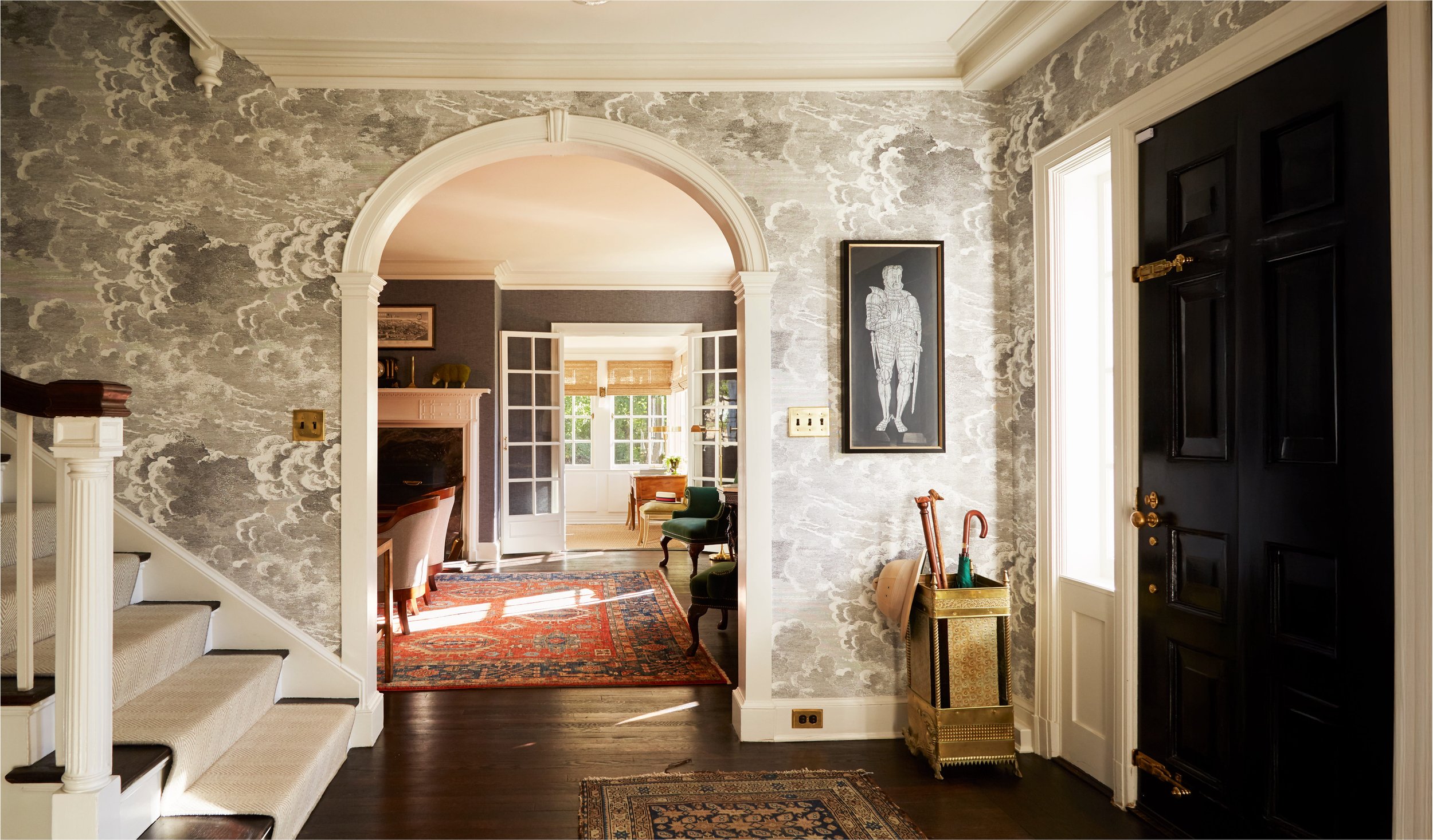 Interior view of a traditional home with patterned wallpaper, a black front door, stairs on the left, and a doorway leading to a bright room with windows and furniture. There are rugs on the floor and paintings on the wall.